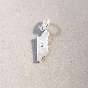 May include: A silver charm in the shape of Belize, with the country's name engraved on the front. The charm has a small loop at the top for attaching to a bracelet or necklace. The charm is on a gray surface.