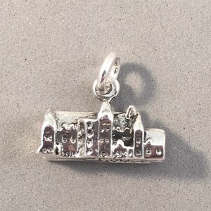 May include: A silver-colored charm depicting a detailed cityscape with multiple buildings and towers. The charm has a loop at the top for attachment to a necklace or bracelet. The charm is made of a shiny, reflective metal.