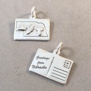 May include: Two silver charms. One charm depicts the state of Nebraska with a bison and a landscape. The other charm is a postcard with the words "Greetings from Nebraska" and a stamp.
