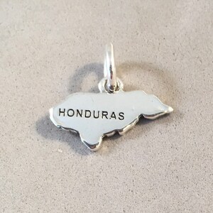 May include: A silver charm in the shape of Honduras, with the country's name in capital letters. The charm has a loop at the top for attaching to a bracelet or necklace. The charm is on a gray surface.