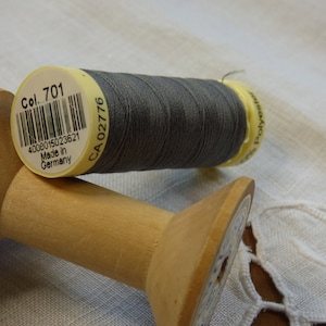 Könnte beinhalten: Eine Spule grauen Fadens auf einer Holzspule. Das Fadenlabel lautet "Col. 701", "4008015023621", "Made in Germany" und "CA 02776".