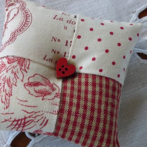 Peut inclure: Un petit coussin à épingles carré avec un motif patchwork. Il présente des sections de tissu crème avec des motifs floraux et à pois rouges, et un motif à carreaux rouges et blancs. Un bouton rouge en forme de cœur fixe les couches.