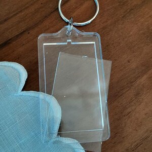Pode incluir: Chaveiro de acr&iacute;lico transparente com um inserto retangular, preso a um chaveiro de metal prateado. O chaveiro tem uma pel&iacute;cula protetora transparente. Um tecido azul claro com borda ondulada &eacute; vis&iacute;vel no canto.