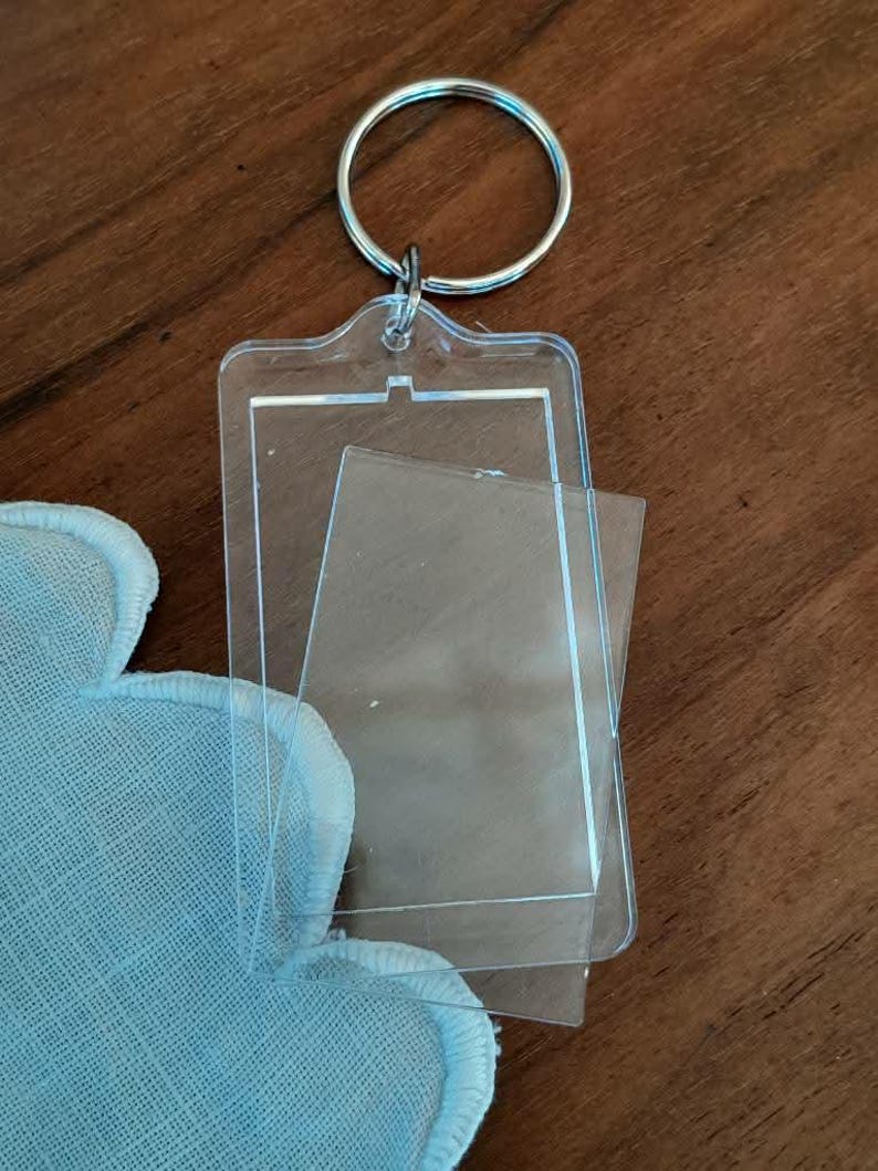 Pode incluir: Chaveiro de acr&iacute;lico transparente com um inserto retangular, preso a um chaveiro de metal prateado. O chaveiro tem uma pel&iacute;cula protetora transparente. Um tecido azul claro com borda ondulada &eacute; vis&iacute;vel no canto.