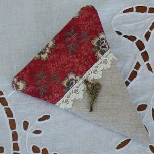 Peut inclure: Un étui à aiguilles triangulaire en tissu avec un imprimé floral rouge et une base en lin beige. Une bordure en dentelle décorative sépare les deux tissus. Une petite paire de ciseaux ornée est attachée à l'étui.