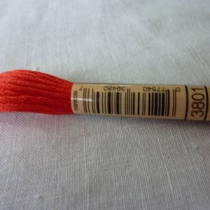 Op de afbeelding: Een felrode borduurgarenspoel met een bruin papieren etiket. Het etiket bevat een barcode, het nummer 3801 en andere gedrukte details. De draad is strak opgerold, tegen een lichtgekleurde stoffen achtergrond.