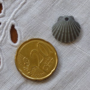 Puede incluir: Una moneda de 20 céntimos de euro de color dorado y una concha de vieira gris sobre un mantel de ganchillo blanco.