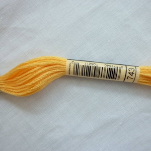 Op de afbeelding: Een klosje DMC borduurgaren in kleur 743, een helder geel. Het garen is gewikkeld om een kartonnen spoel met een wit label waarop het DMC-logo, het kleurnummer en de barcode staan.