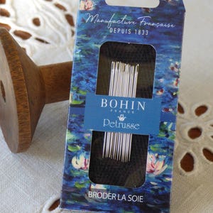 Peut inclure: Une bobine en bois et une boîte d'aiguilles Bohin France. La boîte présente un motif floral bleu et blanc avec le texte "Manufacture Française Depuis 1833" et "BRODER LA SOIE". Les aiguilles sont argentées.