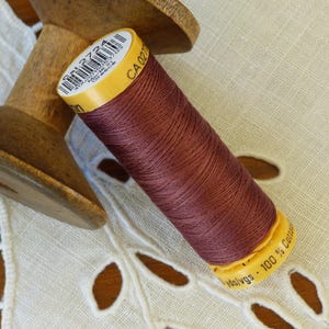 May include: A spool of maroon thread wound on a wooden bobbin. The thread is labeled "CA 0271" and "100% Cotton".