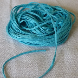 Puede incluir: Una madeja de hilo de bordar azul turquesa, un tipo de hilo utilizado para el bordado a mano, se muestra sobre una superficie de lino blanco. La madeja está enrollada de forma suelta y tiene un hilo largo que se extiende desde la madeja.