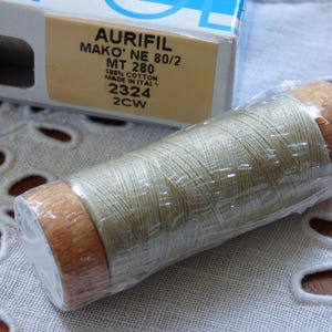 Puede incluir: Un carrete de hilo Aurifil Mako' NE 80/2, MT 280, 100% algodón, de color verde claro. El carrete tiene extremos de madera y está envuelto en plástico transparente. La caja del fondo tiene el texto "AURIFIL".