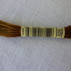 Op de afbeelding: Een bruin borduurgarenklosje met een geel label waarop "829" staat en een barcode. Het label heeft ook de tekst "5433039P".