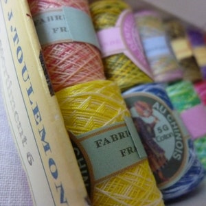 Può includere: Una collezione di rocchetti di filo colorati, tra cui giallo, rosa, verde e blu, con etichette che riportano "Fabriqué en France" e "Au Chinois". I rocchetti sono ordinatamente disposti in una scatola, suggerendo un tema artigianale o di cucito.