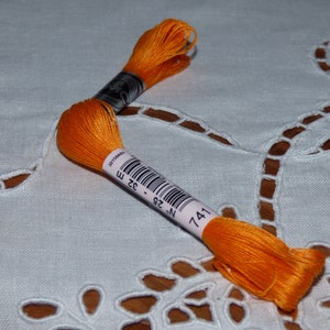 Peut inclure: Fil à broder orange vif, portant le numéro 741, présenté sur un tissu blanc à motifs. Le fil est enroulé sur une bobine avec un code-barres et des informations de mesure. L'image met en valeur la couleur vibrante et la texture du fil.