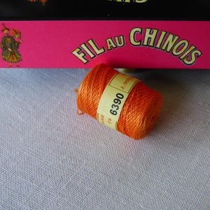 Può includere: Un'bobina di filo da ricamo arancione con il testo "FIL AU CHINOIS" e "6390" sull'etichetta.