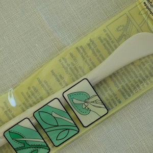 Peut inclure: Un outil d'appliqué en plastique blanc dans un emballage transparent. L'outil a une extrémité incurvée en forme de spatule. L'emballage comporte des illustrations de feuilles vertes et blanches et le texte "useful for marking and cr...applique, reverse applique."