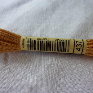 Op de afbeelding: Close-up van een spoel goudbruin borduurgaren. Het etiket toont nummer 437 en een barcode. De draad is strak opgerold, met losse strengen die zich uitstrekken. De achtergrond is een effen, lichtgekleurde stof.