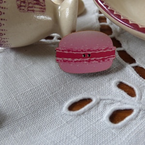 May include: A pink macaron-shaped button with a red stripe and two black button holes. The button is on a white, textured surface with cut-out leaf designs. A cream-coloured teacup and a plate with a red rim are in the background.