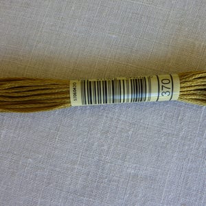 Op de afbeelding: Een streng goudgeel borduurgaren, gemarkeerd met nummer 370. Het garen is strak opgerold en bijeengehouden door een papieren band met een barcode en het nummer 370. De achtergrond is een lichte, getextureerde stof.