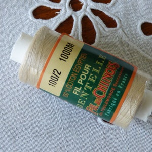 May include: A spool of 100/2 1000M thread made from 100% Egyptian cotton. The label reads "Fil pour dentelle Fil Chinois" and is green and orange. The thread is beige.