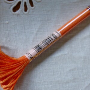 May include: Close-up of a skein of orange embroidery floss. The floss is wrapped around a cardboard tube with a barcode and the number 4124. The background is a white, textured fabric.