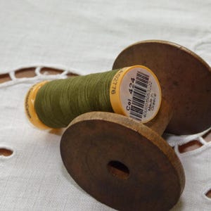 Puede incluir: Una bobina de madera con hilo verde oliva y una etiqueta amarilla con la inscripción "Col. 424". La bobina descansa sobre una tela blanca estampada. El hilo está bien enrollado. La etiqueta también tiene un código de barras.