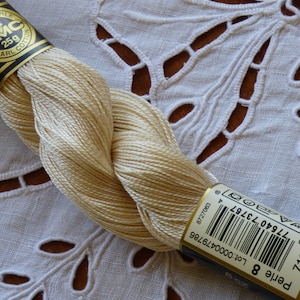 Peut inclure: Une pelote de fil à broder DMC Pearl Cotton de couleur crème. Le fil est enroulé autour d'une étiquette bleu et or avec le logo DMC et le texte "Perle 8 Lot:0000479786 712 8727063 077540 73757 4".