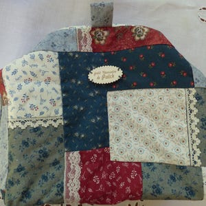 Peut inclure: Un protège-thé en patchwork avec une poignée, composé de tissus à motifs floraux et variés dans les tons de bleu, rouge et crème. Le protège-thé comprend une bordure en dentelle et une petite étiquette en bois avec "petit Nécessaire de Patch".
