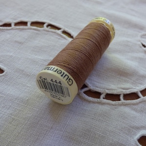 May include: A spool of brown thread, labeled "Gutermann Col. 444," rests on a white, embroidered fabric. The thread is tightly wound, and the label includes a barcode and text indicating it was made in Germany. The fabric has a cut-out design.