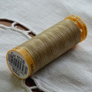 May include: A spool of tan-colored thread with a yellow plastic end cap. The label reads "Col. 1026" and includes a barcode and text indicating it was made in Greece. The thread is tightly wound on the spool.