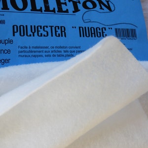 Peut inclure: Un gros plan sur un rouleau de ouate de polyester blanc étiqueté "Polyester "Nuage"" avec le texte "Fabriqué en France" et "61.100.1000" sur l'étiquette bleue. La ouate est décrite comme "Souple", "Mince" et "Léger".