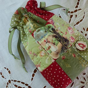 Peut inclure: Une pochette à cordon matelassée avec un motif patchwork de tissus vert sauge et rouge foncé à pois blancs et motifs floraux. Un panneau central représente une silhouette dans un paysage, et un petit bouton en bois montre un personnage à capuche rouge.