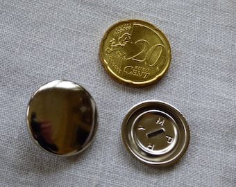 Puede incluir: Dos botones de metal plateado, uno con superficie lisa y el otro con un borde elevado y las letras "YMP" inscritas en el interior. También hay una moneda de 20 céntimos de euro en la imagen.
