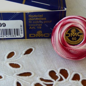 Puede incluir: Una madeja de hilo de algodón para ganchillo DMC 80 en tonos rojos y blancos, con una etiqueta dorada y negra. El hilo está junto a una caja azul con texto en francés y un código de barras. Para ganchillo.