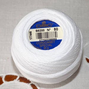 Peut inclure: Une pelote de fil blanc DMC Cordonnet Special, taille 80, avec une étiquette bleue et blanche. L'étiquette indique que le fil pèse 20 g. Le fil est bien enroulé, montrant la texture du coton. Idéal pour le crochet et la broderie.