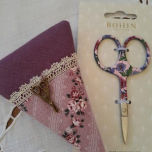 Peut inclure: Une paire de ciseaux fantaisie à motifs floraux, présentée avec un étui en tissu assorti. Les ciseaux sont sur une carte qui indique "BOHIN" et "CISEAUX FANTAISIE FANCY SCISSORS". L'étui est violet et rose avec une bordure en dentelle.