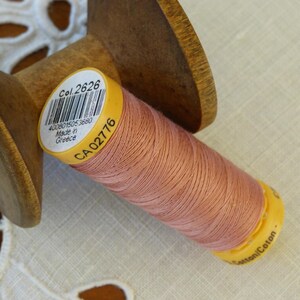 Può includere: Un'bobina di filo di cotone rosa pallido con un'etichetta gialla che recita "Col. 2626 4003801505 32880 Made in Greece CA 02776" e "Cotonni Coton".