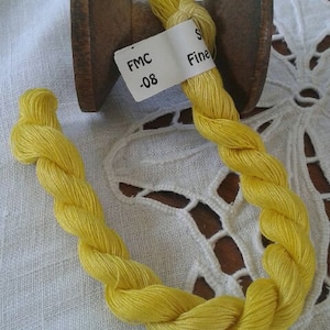 May include: A wooden spool with bright yellow thread, labeled "FMC -08 Fine." The thread is twisted into a decorative shape, with a portion wrapped around the spool. The background is a white, textured surface.