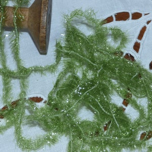 Peut inclure: Une bobine en bois avec de la laine verte, scintillante et duveteuse. La laine est de couleur vert clair et a un aspect scintillant. La laine est partiellement enroulée sur la bobine et partiellement étalée sur une surface blanche brodée.