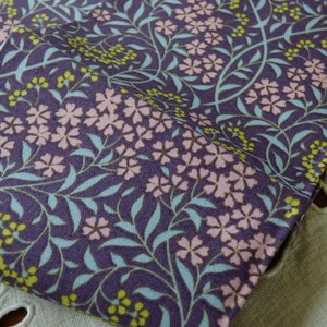 May include: Close-up of a folded fabric with a floral pattern. The fabric features a deep purple background with a repeating design of pink flowers, light blue leaves, and yellow accents. The fabric is likely cotton or a similar material.