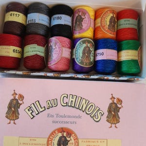 Può includere: Una scatola di 10 rocchetti di filo da ricamo in vari colori, tra cui rosso, giallo, verde, blu e nero. I rocchetti sono etichettati con il nome del marchio "Fil au Chinois" e il nome dell'azienda "Ets Toulemonde successeurs".
