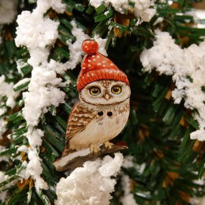 Peut inclure: Ornement en bois en forme de hibou portant un bonnet rouge à pois blancs. Le hibou a de grands yeux jaunes et est perché sur une branche. L'ornement est placé devant un sapin de Noël enneigé.