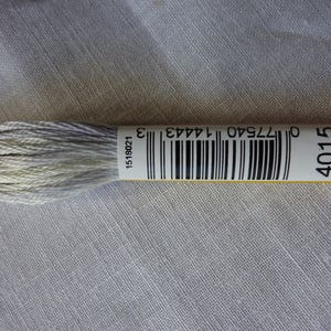 Könnte beinhalten: Ein Strang hellgraues Stickgarn mit einem Barcode-Etikett. Das Etikett zeigt die Nummer 4015 und einen Barcode. Das Garn ist fest gewickelt und wirkt weich. Der Hintergrund ist ein hellgrauer Stoff.