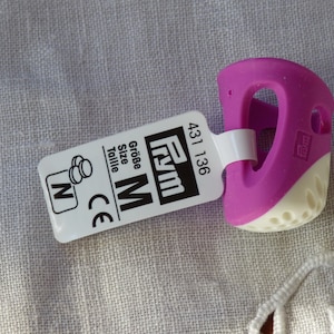 Peut inclure: Un protège-doigt en silicone violet et blanc avec une étiquette indiquant "Frym Taille M Grosse Taille CE 431 136".