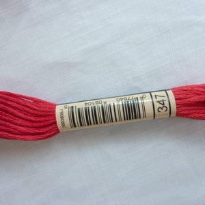 Op de afbeelding: Een spoel rood borduurgaren, gemarkeerd met nummer 347, ligt op een witte ondergrond. Het etiket bevat een barcode en andere nummers. De draad is strak opgerold, wat de levendige kleur benadrukt.