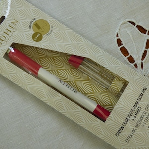 Peut inclure: Un crayon craie porte-mine extra fin Bohin, blanc et rouge, avec 6 mines, présenté dans un emballage crème et or. L'emballage porte le texte "BOHIN" et "0,9 mm".