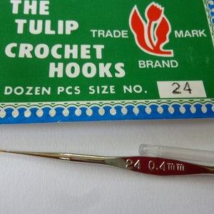 Puede incluir: Un ganchillo del número 24 con una punta metálica fina y una funda de plástico transparente. El embalaje, con fondo verde, muestra el texto "THE TULIP CROCHET HOOKS" y un logotipo de tulipán rojo. El ganchillo tiene 0,4 mm de diámetro.