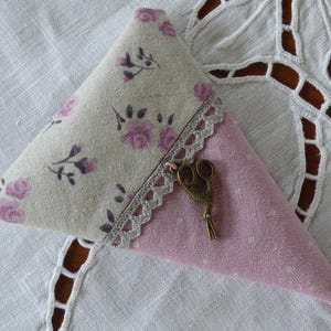 Puede incluir: Un cojín triangular con un estampado floral. La sección superior presenta una tela color crema con rosas rosas, mientras que la inferior es rosa con lunares blancos. Un ribete de encaje decorativo y un pequeño dije de tijeras doradas añaden detalles.