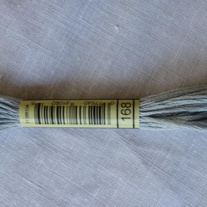 Op de afbeelding: Een streng lichtgrijs borduurgaren met een geel label met de tekst "168". Het garen is strak opgerold en ziet er zacht uit. De achtergrond is een lichtgrijze stof.